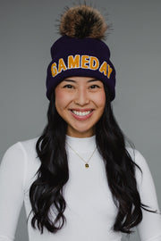 A navy blue ribbed knit beanie features bold "GAMEDAY" lettering in gold chenille across the cuff, topped with a luxurious faux-fur pom-pom in brown and black tones. The classic cuffed design combines spirited collegiate styling with cozy winter functionality, perfect for game day or everyday cold-weather wear.
