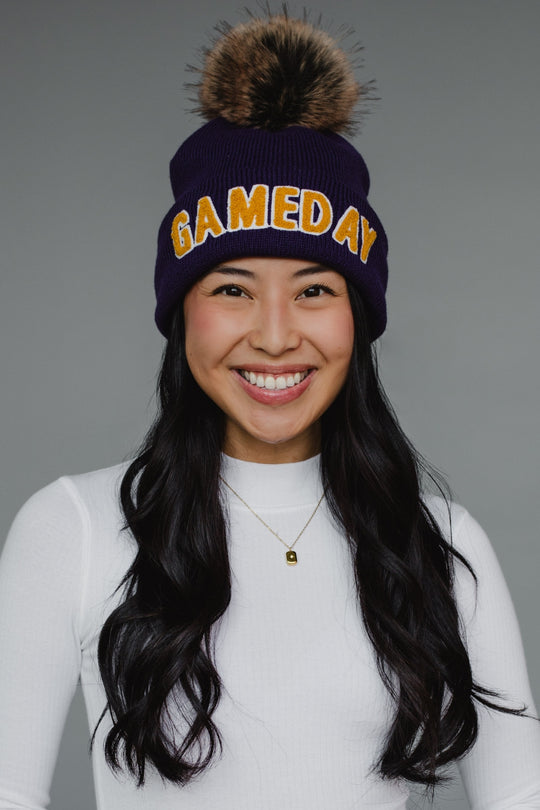 A navy blue ribbed knit beanie features bold "GAMEDAY" lettering in gold chenille across the cuff, topped with a luxurious faux-fur pom-pom in brown and black tones. The classic cuffed design combines spirited collegiate styling with cozy winter functionality, perfect for game day or everyday cold-weather wear.