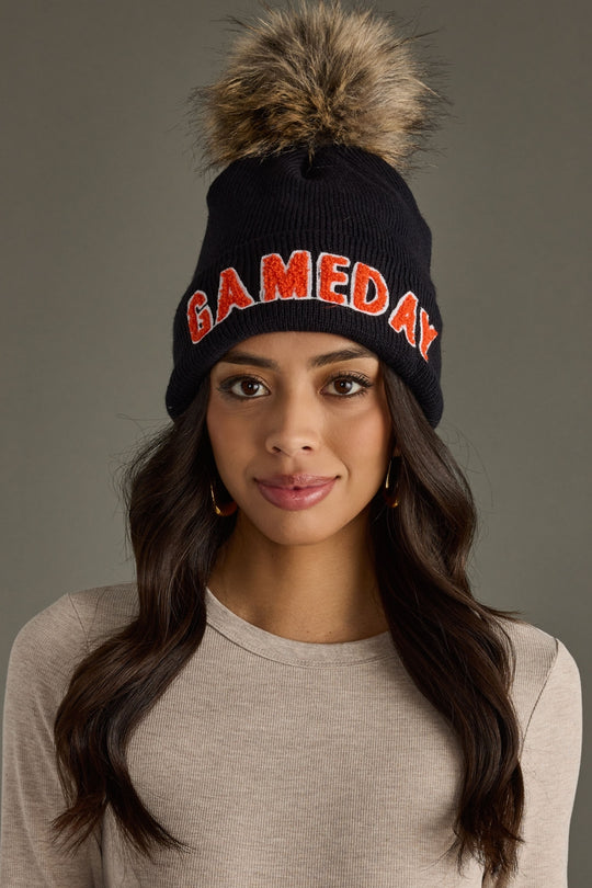 Woman with long dark hair wearing Panache Apparel’s black and orange Game Day pom hat with bold lettering and a faux fur pom.