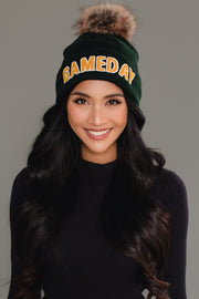 A smiling woman with long dark hair wears a Panache Apparel Collection Green & Gold Game Day Pom Hat, featuring a fleece lining and a fur pom-pom, standing against a plain gray background.