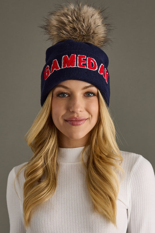 Blonde woman wearing Panache Apparel’s navy and red Game Day pom hat with bold lettering, a ribbed cuff, and faux fur pom.