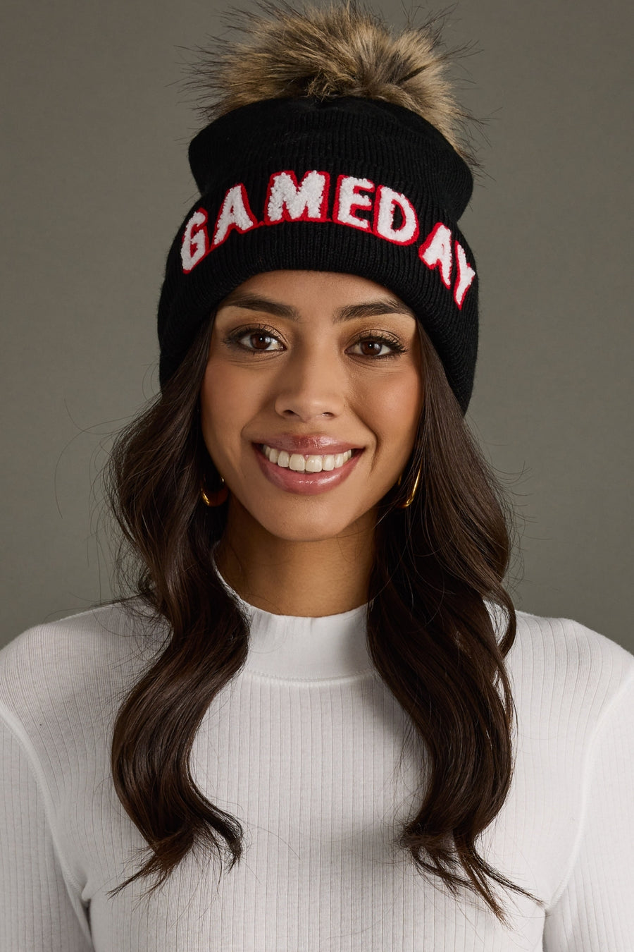 Smiling woman wearing Panache Apparel’s black Game Day pom hat with white and red lettering, a ribbed cuff, and faux fur pom.