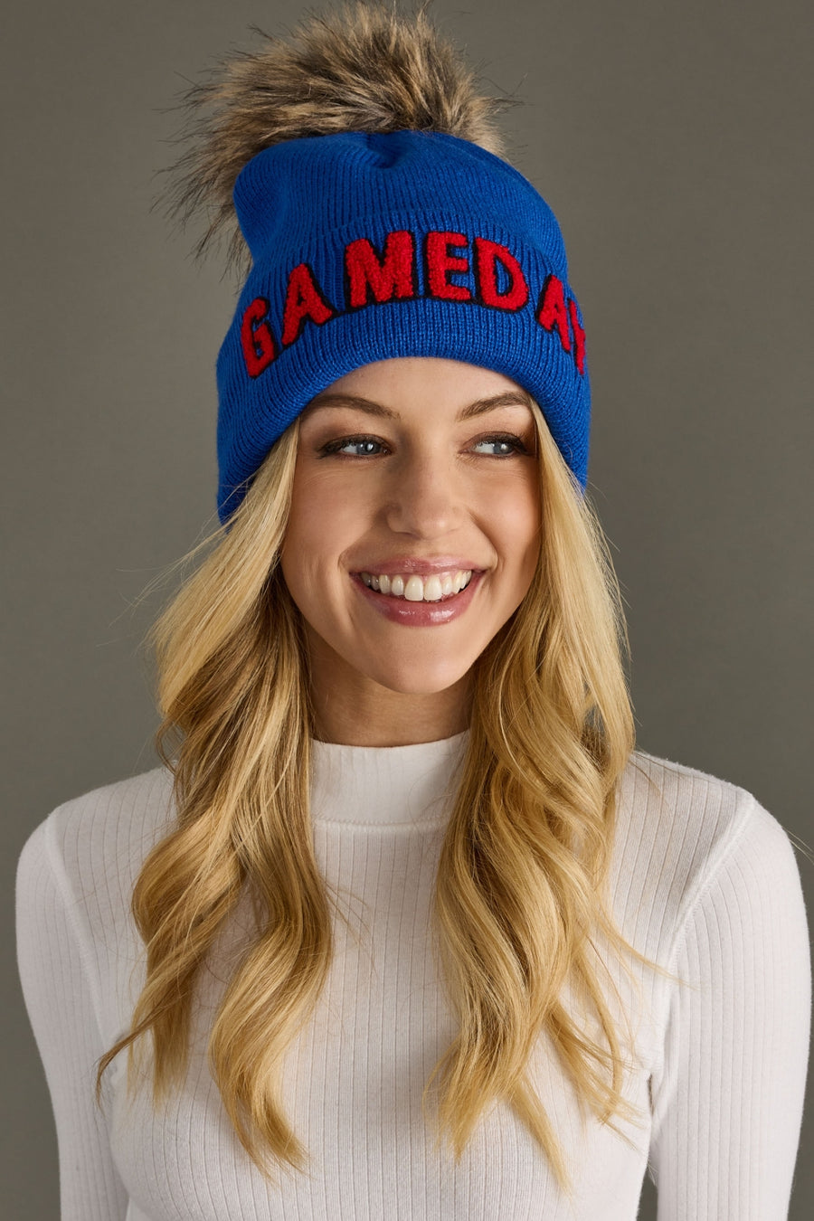 Smiling blonde woman wearing Panache Apparel’s royal blue and red Game Day pom hat with bold lettering and a faux fur topper.