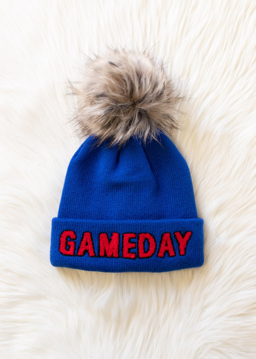 Flat lay of Panache Apparel’s royal blue Game Day pom hat featuring red lettering, a folded cuff, and faux fur detail.