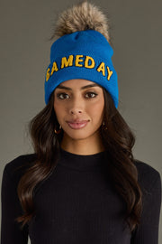 Brunette model wearing Panache Apparel’s blue and gold Game Day pom hat with bold lettering and a faux fur topper.