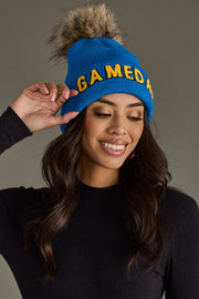 Woman smiling in Panache Apparel’s blue Game Day knit hat with gold text, a folded cuff, and pom detail.