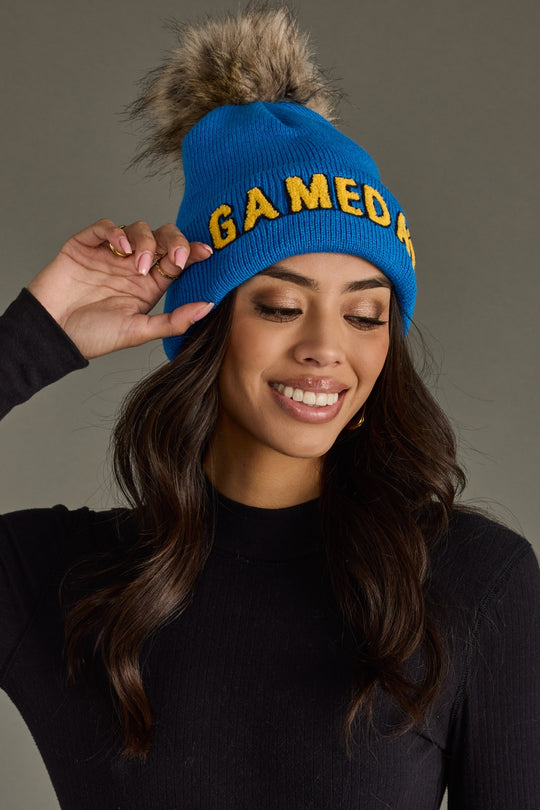 Woman smiling in Panache Apparel’s blue Game Day knit hat with gold text, a folded cuff, and pom detail.