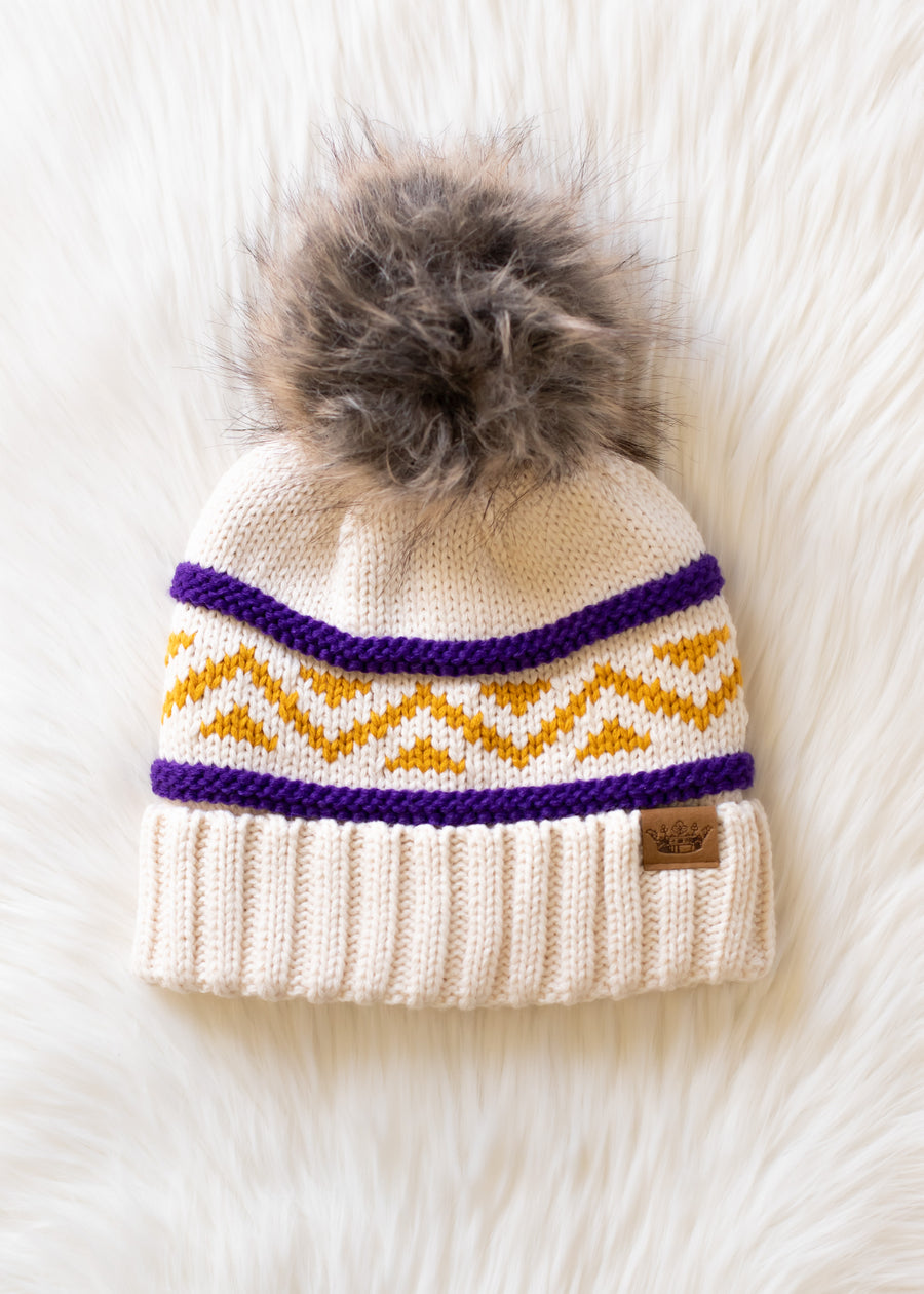 A Cream, Purple & Gold Patterned Pom Hat by Panache Apparel Collection, featuring a cream knit design with a brown faux fur pom-pom, yellow zigzag and purple stripe patterns, displayed on a soft white textured surface.