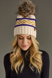 A woman with long blonde hair models the Panache Apparel Collection Cream, Purple & Gold Patterned Pom Hat, featuring a cream knit design with a fur pom-pom, yellow zigzag accents, and purple stripes. She pairs it with a black turtleneck and looks down with a slight smile against a gray background.
