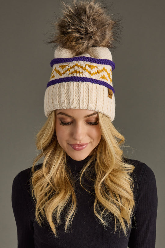 A woman with long blonde hair models the Panache Apparel Collection Cream, Purple & Gold Patterned Pom Hat, featuring a cream knit design with a fur pom-pom, yellow zigzag accents, and purple stripes. She pairs it with a black turtleneck and looks down with a slight smile against a gray background.