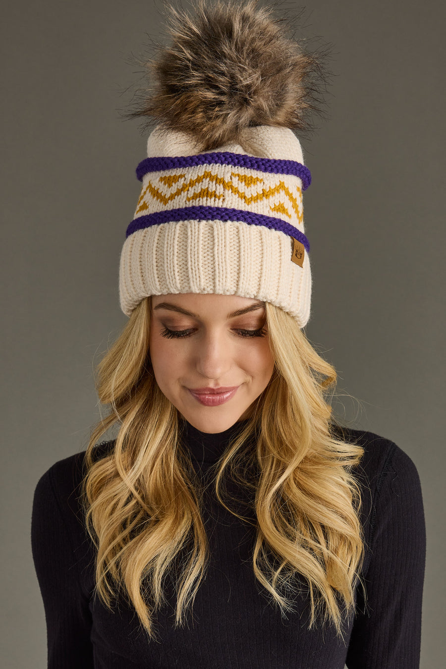 A woman with long blonde hair models the Panache Apparel Collection Cream, Purple & Gold Patterned Pom Hat, featuring a cream knit design with a fur pom-pom, yellow zigzag accents, and purple stripes. She pairs it with a black turtleneck and looks down with a slight smile against a gray background.