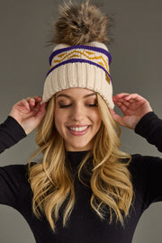 A woman with long blonde hair smiles while adjusting the Panache Apparel Collection Cream, Purple & Gold Patterned Pom Hat, featuring a knit design and pom-pom, paired with a black top against a plain background.