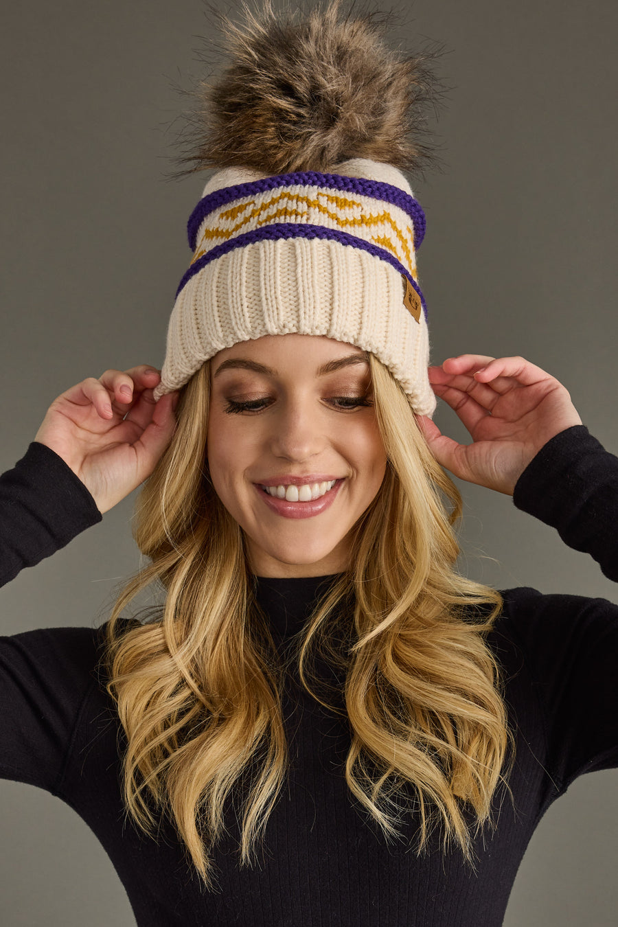 A woman with long blonde hair smiles while adjusting the Panache Apparel Collection Cream, Purple & Gold Patterned Pom Hat, featuring a knit design and pom-pom, paired with a black top against a plain background.