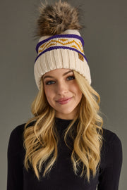 A woman with long blonde hair wears the Panache Apparel Collection Cream, Purple & Gold Patterned Pom Hat, featuring a large faux fur pom-pom. She smiles slightly and wears a black top against a plain gray background.