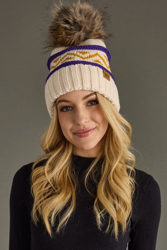 A woman with long blonde hair wears the Panache Apparel Collection Cream, Purple & Gold Patterned Pom Hat, featuring a large faux fur pom-pom. She smiles slightly and wears a black top against a plain gray background.
