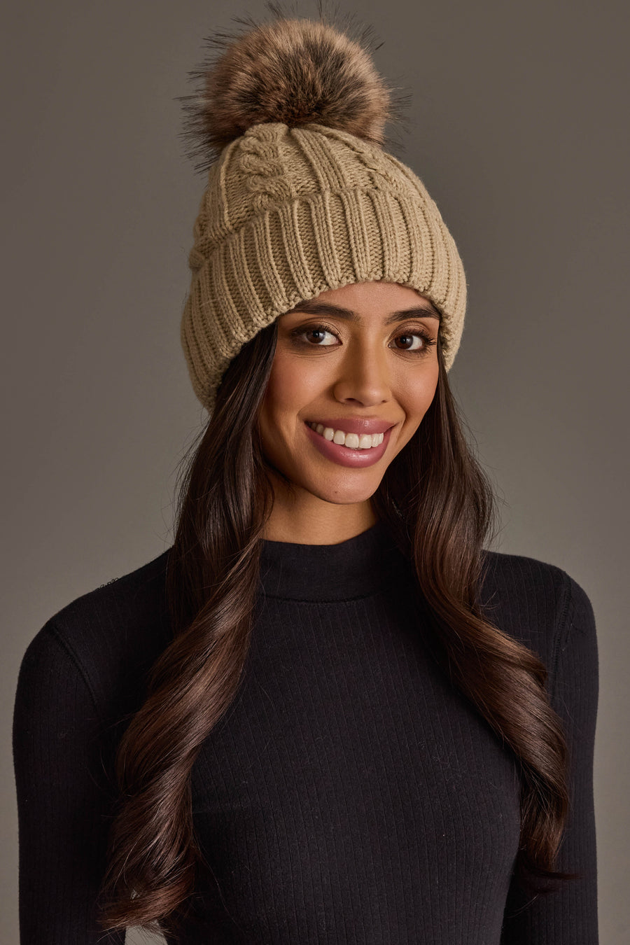 Smiling woman with long brown hair wearing the Panache Apparel Collection Tan Cable Knit Design Pom Hat and a black ribbed turtleneck sweater, against a neutral background.