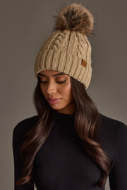 A woman with long brown hair wears the Panache Apparel Collection Tan Cable Knit Design Pom Hat, styled with a fitted black sweater, as she looks down against a neutral background.