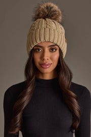 A woman with long, wavy brown hair faces the camera against a plain background, wearing the Panache Apparel Collection’s Tan Cable Knit Design Pom Hat with a faux fur pom-pom and a black ribbed turtleneck sweater.