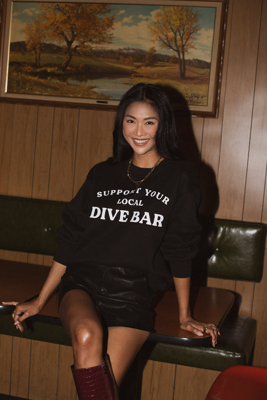 A woman sits on a booth edge in a wood-paneled room, smiling in the black Panache Apparel Collection 'Support Your Local Dive Bar' oversized sweatshirt, black shorts, and red boots. A landscape painting hangs on the wall behind her.