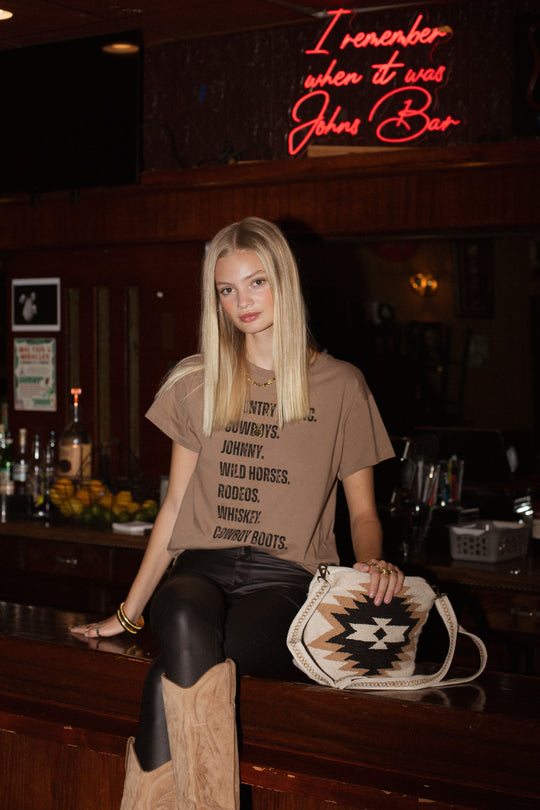A young woman with long blonde hair stands at a rustic bar, wearing Panache Apparel Collection’s Western List Tee, made from 100% cotton, paired with black pants, tan cowboy boots, and holding a patterned bag by a red neon sign.