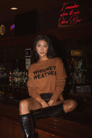 A woman with long dark hair sits on a bar counter wearing the Panache Apparel Collection's Whiskey Weather Sweater in brown and black knee-high boots. Glowing neon lights and rows of bottles behind her create the perfect cozy vibe.