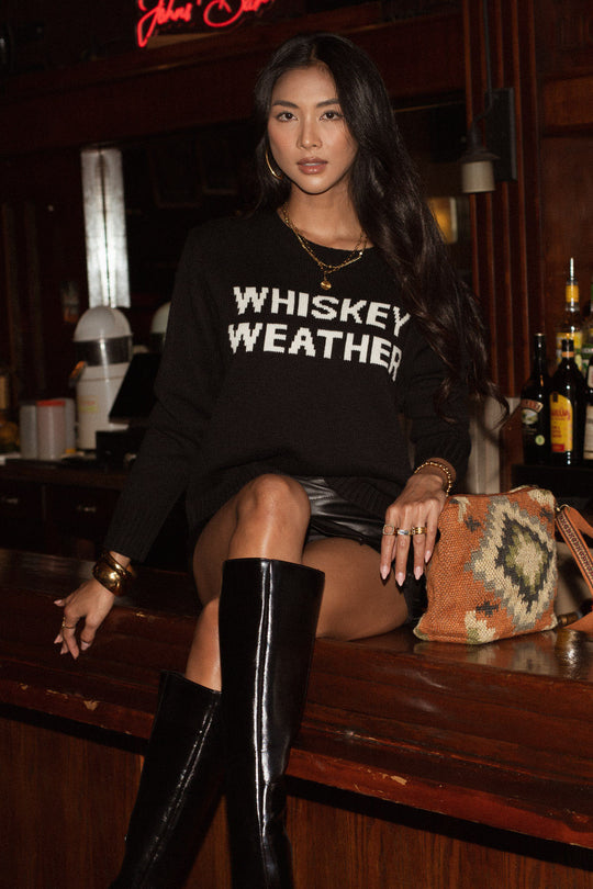 A woman with long dark hair sits on a bar counter wearing the Panache Apparel Collection Whiskey Weather Sweater - Black, paired with a black leather skirt and tall boots. Bottles and bar equipment are visible in the background.