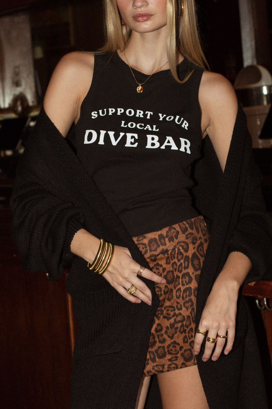 A woman wears the Panache Apparel Collection's Support Your Local Dive Bar Tank Top with a leopard-print skirt, gold rings and bangles, and a black cardigan draped over her arms.