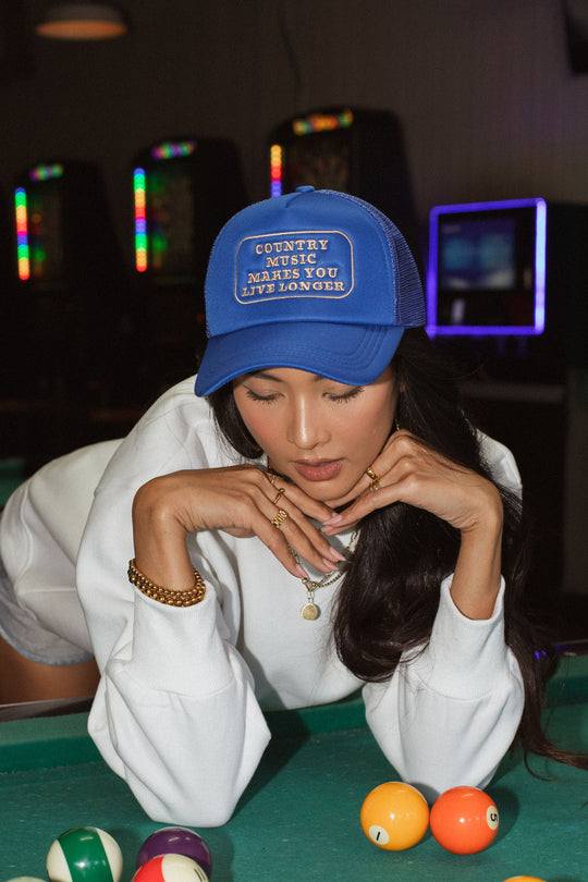 A woman wearing the Panache Apparel Collection Country Music Trucker Hat - Blue, which reads "Country Music Makes You Live Longer," leans over a pool table in a white sweatshirt and gold jewelry, with pool balls in the foreground.