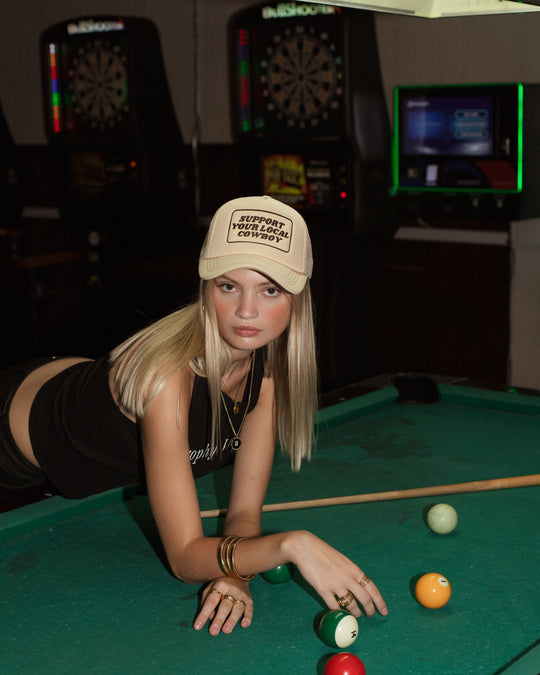 A young woman with long blonde hair leans over a pool table in a black tank top and gold jewelry, wearing Panache Apparel Collection’s Support Your Local Cowboy Trucker Hat with an adjustable snapback. Dart machines are visible in the background.
