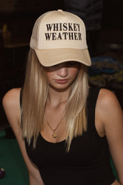 A woman with long blonde hair wears the Panache Apparel Collection Whiskey Weather Trucker Hat - Tan & Brown, featuring bold lettering. Paired with a black sleeveless top and gold necklace, she looks downward. The snapback ensures comfort.