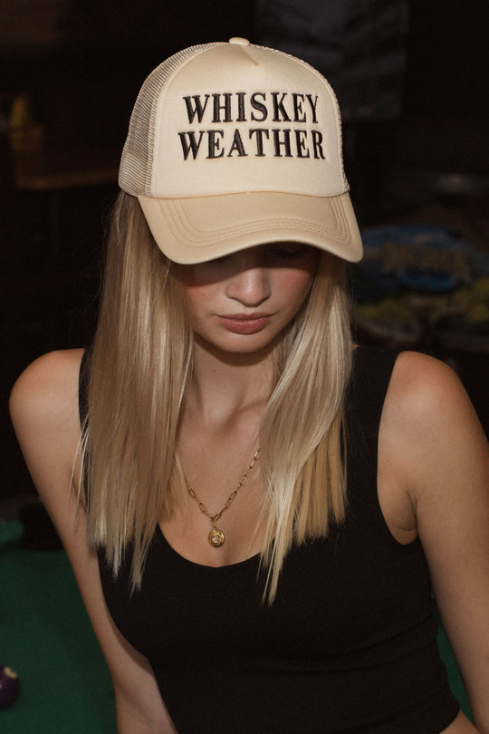 A woman with long blonde hair wears the Panache Apparel Collection Whiskey Weather Trucker Hat - Tan & Brown, featuring bold lettering. Paired with a black sleeveless top and gold necklace, she looks downward. The snapback ensures comfort.