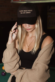 A woman with long blonde hair wears the Panache Apparel Collection Country Music Trucker Hat in black, paired with a black tank top, beige cardigan, and gold jewelry as she stands in front of a pool table.
