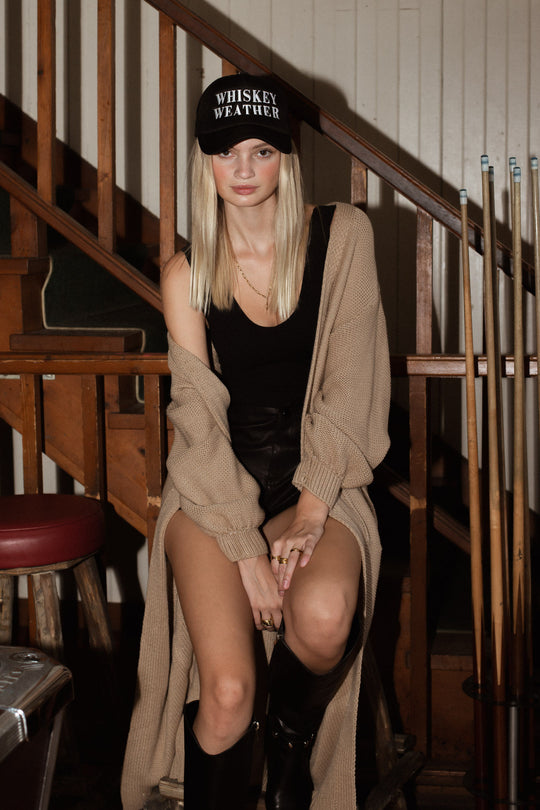 A woman sits indoors on a bench near pool cues, wearing the tan Hilton Duster Cardigan from Panache Apparel Collection over a black cap, tank top, shorts, and knee-high boots, looking into the camera with a neutral expression.