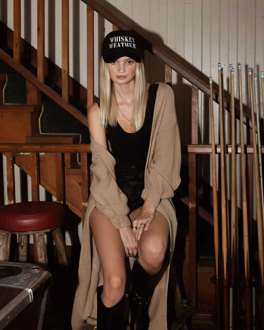 A woman in a black hat, black top, shorts, and tall boots sits by a pool table and staircase, layering her outfit with the Hilton Duster Cardigan - Tan from Panache Apparel Collection. Pool cues hang on the wall behind her.