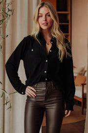 A woman with long blonde hair stands indoors, smiling softly with a hand on her hip, wearing the Panache Apparel Collection Black Super Soft Button Up and high-waisted brown leather pants.