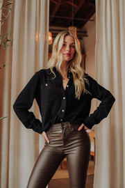 A woman with long blonde hair poses confidently indoors in front of beige curtains, wearing the Panache Apparel Collection Black Super Soft Button Up tucked into high-waisted shiny brown pants, her hands resting on her hips.