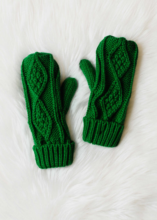 The Highland Mittens by panacheaccessories are green cable knit mittens with a diamond pattern, shown on a white fluffy surface.