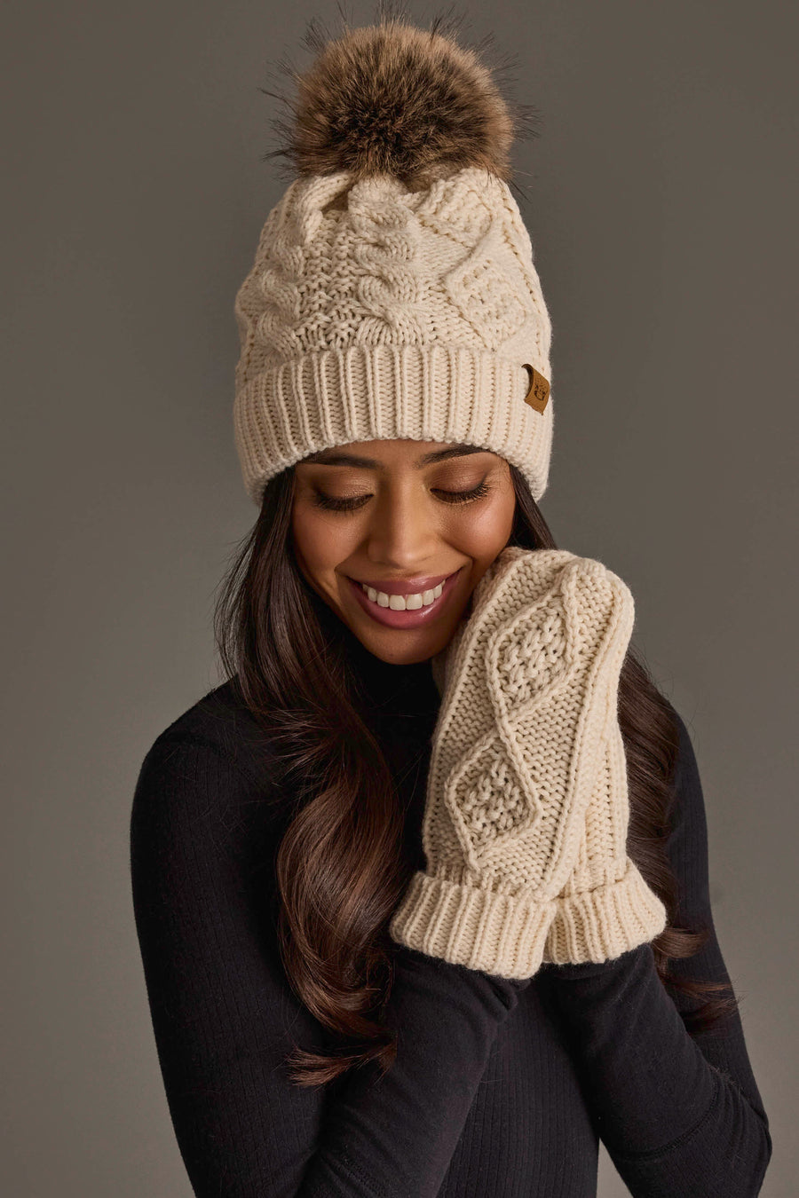 A woman smiles while wearing the Panache Apparel Collection Ivory Cable Knit Pom Hat and matching fleece-lined mittens, set against a plain gray background.
