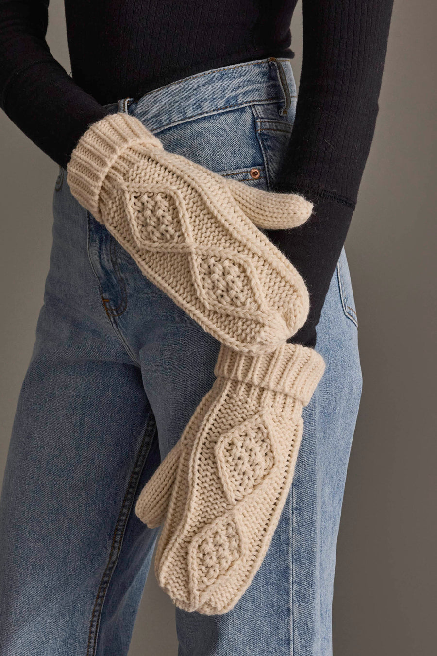 A person wearing a black long-sleeve shirt and blue jeans has their arms crossed, showing off the cozy Ivory Cable Knit Mittens from Panache Apparel Collection.