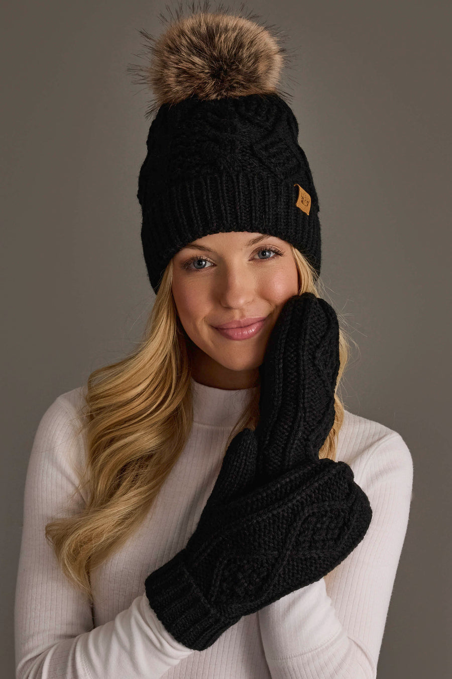 A smiling woman in a white sweater wears the Panache Apparel Collection’s Black Cable Knit Pom Hat, featuring a fleece-lined interior and faux fur pom, along with matching black mittens. She holds one mitten to her face against a neutral background.