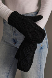 A person in a white ribbed sweater and blue jeans clasps their hands, highlighting the Panache Apparel Collection Black Cable Knit Mittens, featuring a fleece lining and diamond pattern.