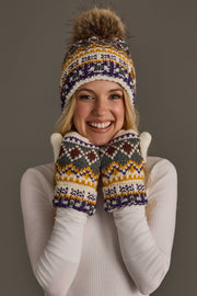 A smiling person wearing the Panache Apparel Collection Ivory, Purple & Gold Patterned Pom Hat with a faux fur pom-pom and matching fleece-lined mittens holds their hands to their face. The hat and mittens feature yellow, purple, and white designs.
