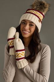 A woman smiles while wearing the Panache Apparel Collection’s Cream, Burgundy & Gold Patterned Pom Hat, a fleece-lined style topped with a faux fur pom, along with coordinating mittens, all set against a plain gray background.