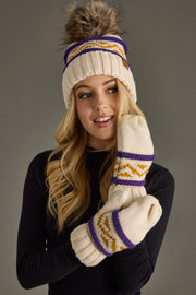 A woman wearing the Panache Apparel Collection’s Cream, Purple & Gold Patterned Pom Hat poses against a gray background, smiling softly.