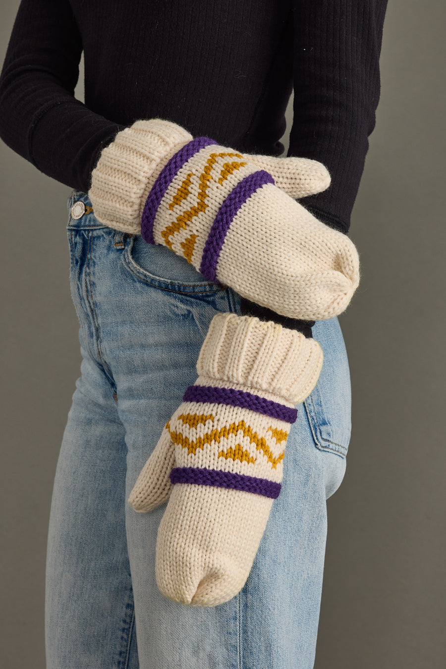 A person wears the Panache Apparel Collection Cream, Purple & Gold Patterned Mittens, paired with blue jeans and a black ribbed sweater, standing against a plain gray background.