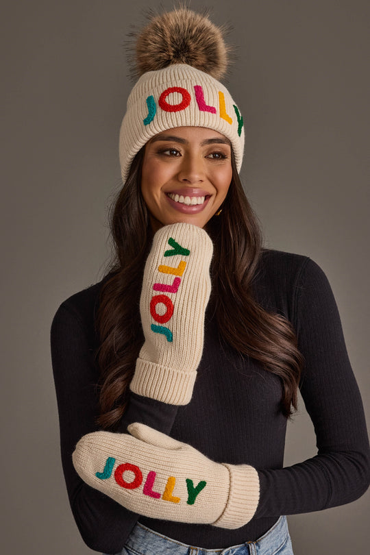 A woman wears a Beige Jolly Pom Hat and matching mittens, both featuring the word JOLLY in colorful embroidered letters.