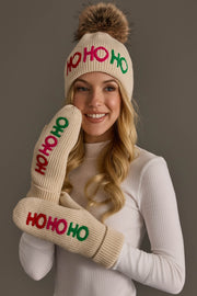 A person wears a Beige Ho Ho Ho Pom Hat and matching mittens, both featuring repeating red and green embroidered HO HO HO text.