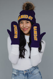 A smiling person wearing the Panache Apparel Collection’s Purple & Gold Game Day Pom Hat and matching fleece-lined mittens, paired with a white long sleeve shirt and blue jeans, poses against a plain gray background.