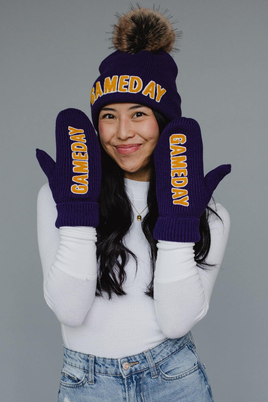 A smiling person wearing the Panache Apparel Collection’s Purple & Gold Game Day Pom Hat and matching fleece-lined mittens, paired with a white long sleeve shirt and blue jeans, poses against a plain gray background.