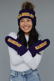 A smiling person wears the Panache Apparel Collection’s Purple & Gold Game Day Pom Hat—featuring yellow "GAMEDAY" lettering—paired with matching mittens, a white top, and jeans, arms crossed against a gray background. The fleece-lined hat offers extra warmth and cozy comfort.
